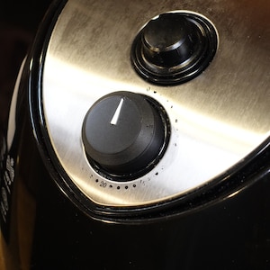 May include: Close-up of a black air fryer with a stainless steel control panel. The control panel has two knobs, one for temperature and one for time. The knobs are black with white markings.