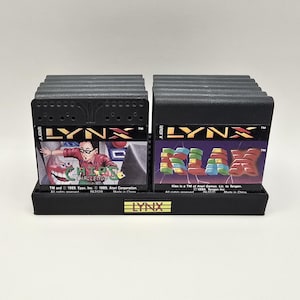 May include: A black plastic display stand holding two rows of Atari Lynx video game cartridges. The left cartridge is labeled Chip's Challenge and the right cartridge is labeled Klax. The base of the stand features a small gold-colored Lynx logo.