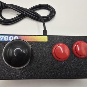 May include: A black rectangular video game controller featuring a large black spherical joystick on the left and two circular red buttons on the right. A label with the text 7800 and a multi-colored gradient bar is positioned above the joystick.