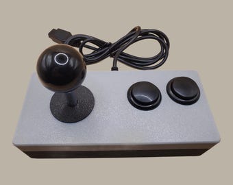 May include: A rectangular arcade-style game controller featuring a light gray top panel, a black ball-top joystick, and two black circular push buttons. A black wired cable is coiled behind the controller, which has a dark gray base.