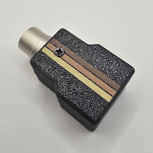 May include: A black rectangular electronic connector featuring a textured surface with two diagonal stripes in yellow and brown. A single black Phillips head screw is positioned near the top edge, and a metallic cylindrical port extends from the side.