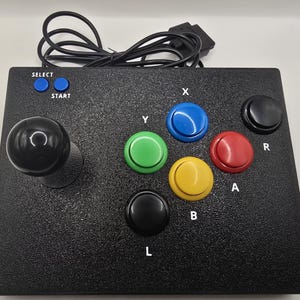 Puede incluir: Un mando de juego estilo arcade de color negro con un joystick negro grande a la izquierda, dos botones azules pequeños etiquetados como SELECT y START, y seis botones de acción de colores más grandes etiquetados como X, Y, A, B, L y R.