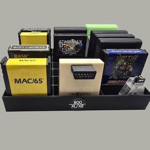 May include: A black plastic storage tray labeled 800 XL/XE containing various rectangular video game cartridges. The collection includes yellow MAC/65 cartridges, a beige FujiNet device, a black AtariMax cartridge, and a blue Miner 2049er game.