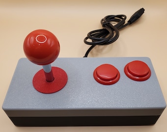May include: A rectangular arcade-style game controller featuring a gray top surface, a black base, and a red ball-top joystick. The controller includes two round red action buttons and a black wired cable extending from the back of the unit.