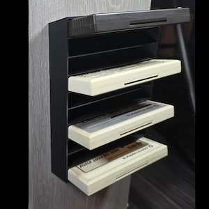 May include: Black plastic cartridge holder with four beige game cartridges. The cartridges are labeled with text. The holder is mounted on a light wood grain surface. The top of the holder has a slot for an additional cartridge.