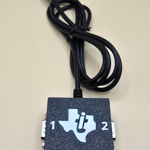 May include: A black 2-port serial splitter with a black cable and a white logo that says "ti" with a stylized Texas shape. The splitter has two DB-9 female connectors labeled "1" and "2".