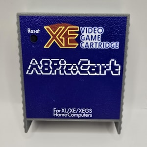 May include: A gray and blue video game cartridge with a textured plastic casing. The label features the text XE VIDEO GAME CARTRIDGE, the word Reset next to a small black button, the stylized text AlphaCart, and For XL/XE/XEGS Home Computers.