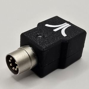 Op de afbeelding: Zwarte adapter met een zilveren connector en het Atari-logo in wit. De adapter heeft een getextureerd oppervlak en een schroef aan de bovenkant. De connector heeft meerdere gouden pinnen.