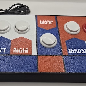 Może przedstawiać: Kontroler do gier w stylu retro z czarną podstawą i kolorowym górnym panelem. Panel podzielony jest na białe, niebieskie i pomarańczowe sekcje, z białymi przyciskami i czerwonym przyciskiem. Etykiety tekstowe to "LEFT", "RIGHT", "WARP", "THRUST" i "FIRE".