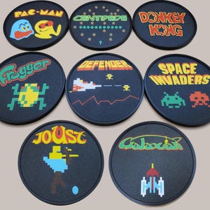 Könnte beinhalten: Acht runde schwarze Untersetzer mit bunten Retro-Videospielgrafiken. Die Untersetzer tragen die Namen "Pac-Man", "Centipede", "Donkey Kong", "Frogger", "Defender", "Space Invaders", "Joust" und "Galaxian".