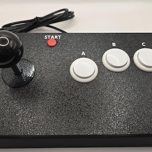 May include: A black rectangular arcade controller featuring a black joystick with a spherical top, a small red button labeled START, and three white circular buttons labeled A, B, and C. A black wired cable with a connector extends from the back.