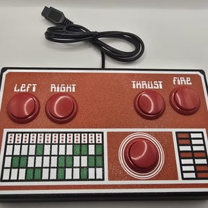 May include: Retro-style game controller with a red and white design. Features four red buttons labeled "LEFT", "RIGHT", "THRUST", and "FIRE". Includes a large central red button and a series of smaller buttons. A black USB cable is attached.