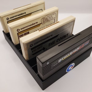 May include: A black plastic storage case for Commodore VIC-20 game cartridges. The case holds four cartridges, with the titles "The Count", "VIC-1917", "VIC-1918", and "Perult 2" visible. The case has a Commodore logo on the front.