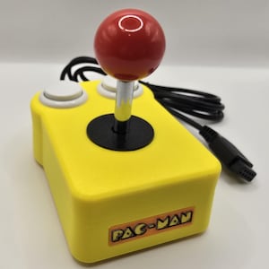 Könnte beinhalten: Ein gelber Pac-Man-Arcade-Joystick mit einem roten, kugelförmigen Griff. Der Joystick hat zwei weiße Knöpfe und eine schwarze Basis. Das Pac-Man-Logo ist auf der Vorderseite sichtbar. Ein schwarzes Kabel und ein Stecker sind angebracht.