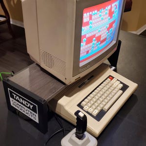 May include: A vintage beige Tandy computer with a monitor displaying a colorful pixelated game, a keyboard, and a black joystick. The monitor sits on a dark gray stand labeled TANDY COMPUTER PRODUCTS in white text on a black background.