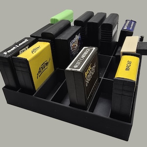 May include: A black plastic grid organizer holding multiple rectangular game cartridges. The cartridges feature labels with text including FastLoad, Action!, Left Cartridge, Basic Computing Language, and MAC/65. Some cartridges are black, yellow, or white.