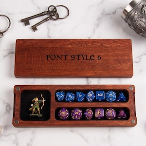 May include: A rectangular wooden box with a lid engraved with the text FONT STYLE 6. The open base contains a miniature archer figure in a black-lined compartment and two rows of polyhedral dice in blue and purple colors, respectively.