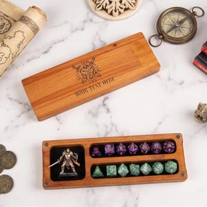 Peut inclure: Boîte à dés en bois avec un compartiment pour dés et une figurine miniature. La boîte est ouverte, révélant un ensemble de dés violets et verts. Un couvercle en bois avec le texte "YOUR TEXT HERE" est sur le dessus. Une carte, une boussole et des livres sont également visibles.