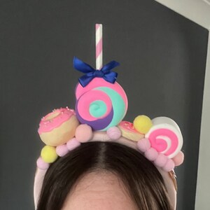 May include: A pink headband with a variety of colorful candy decorations, including a pink and blue swirl lollipop, a pink and white swirl candy, a pink donut with sprinkles, and yellow and pink pom-poms.