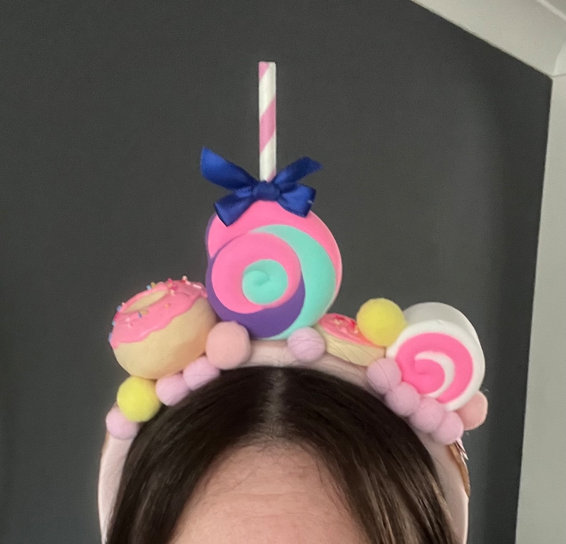 May include: A pink headband with a variety of colorful candy decorations, including a pink and blue swirl lollipop, a pink and white swirl candy, a pink donut with sprinkles, and yellow and pink pom-poms.