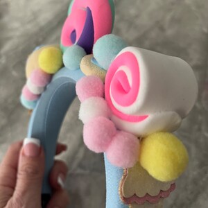 May include: A blue headband with a variety of colorful decorations, including pink and white swirl candies, pastel pom-poms, and a light blue and pink ice cream cone.