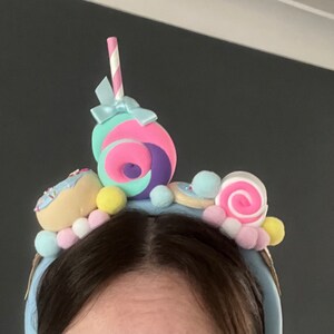 May include: A blue headband with a variety of colorful candy and donut decorations. The headband features a pink and purple swirl candy, a donut with sprinkles, a pink and white swirl candy, and a blue and white striped straw.