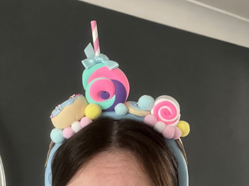 May include: A blue headband with a variety of colorful candy and donut decorations. The headband features a pink and purple swirl candy, a donut with sprinkles, a pink and white swirl candy, and a blue and white striped straw.