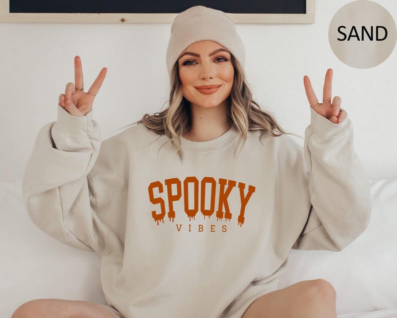 May include: A sand colored sweatshirt with the text 'SPOOKY VIBES' in orange lettering. The sweatshirt has a relaxed fit and a crew neck.