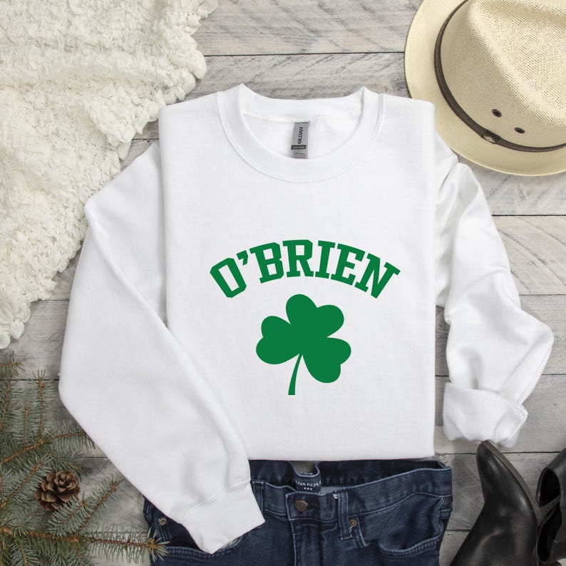 A white long-sleeved shirt with the text "O'BRIEN" printed on it, a pair of jeans, a hat, and some other clothing items are laid out on a wooden surface.