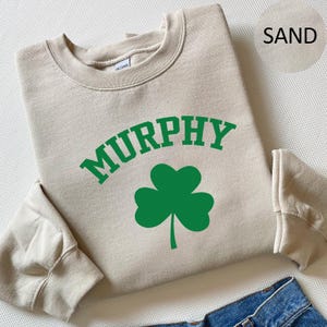A beige sweatshirt with the word "MURPHY" printed on it, along with a green shamrock symbol, is displayed on a surface.
