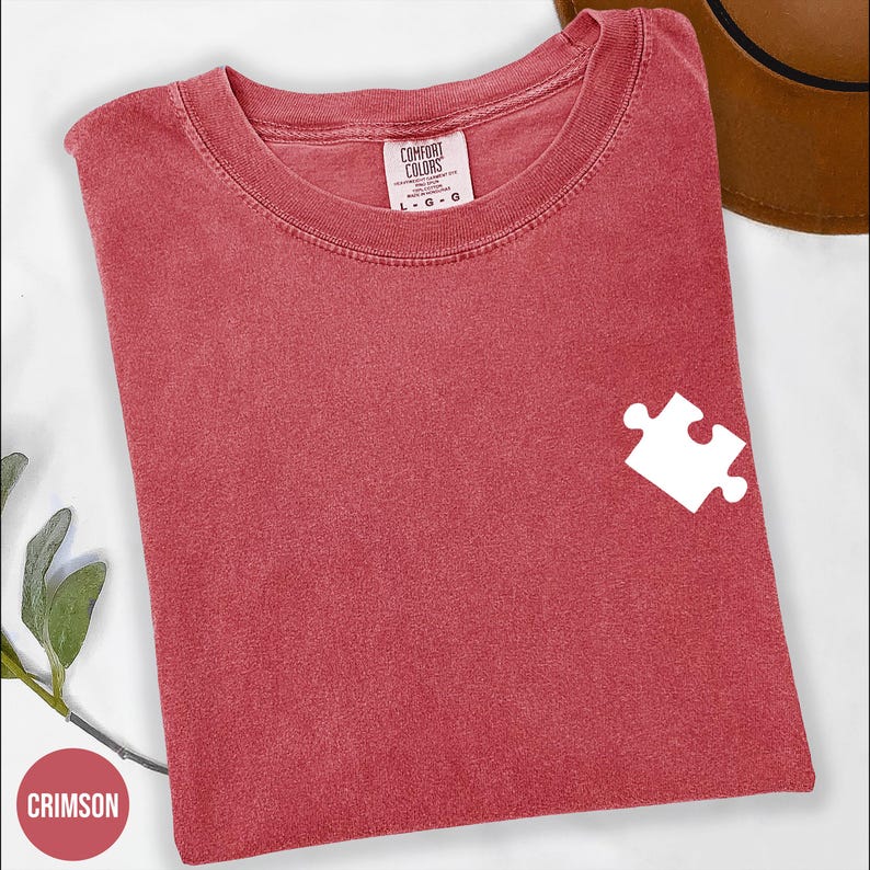 a red t-shirt with a white puzzle piece design on the left side, placed on a white surface next to a brown hat.