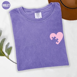 a purple t-shirt with a pink heart-shaped puzzle piece design on the front, placed on a white surface alongside a brown hat.