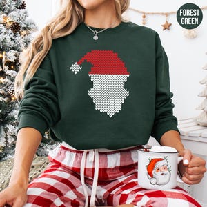 May include: Forest green sweatshirt featuring a knitted Santa Claus design in red and white. The Santa Claus graphic includes a hat and beard. The model is wearing red and white plaid pajama pants and holding a mug with a Santa Claus graphic.