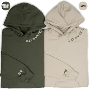 May include: Two hooded sweatshirts, one military green and one sand-colored, are displayed. Both have the Roman numeral "II.XV.MMXXVI" embroidered on the neckline. The military green sweatshirt has a letter "N" and a star on the sleeve, while the sand-colored one has "E S" and a heart.