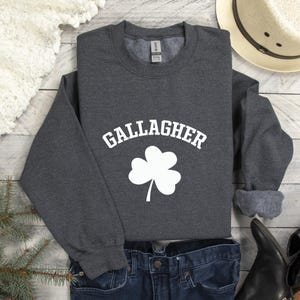 A gray sweatshirt with the word "Gallagher" printed on it, along with a white cloverleaf symbol, is displayed on a wooden surface.