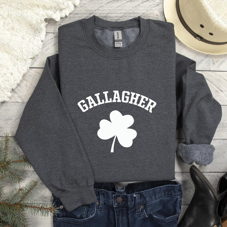 A gray sweatshirt with the word "Gallagher" printed on it, along with a white cloverleaf symbol, is displayed on a wooden surface.