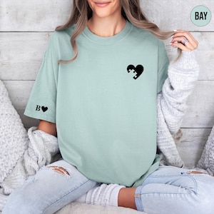 A woman wearing a mint green t-shirt with a black heart-shaped puzzle piece design, sitting on a couch with her hair in a ponytail.