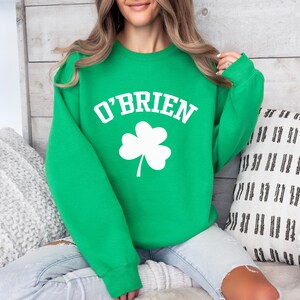 A woman wearing a green sweatshirt with the word "O'Brien" printed on it, sitting on a couch with a white and black patterned pillow behind her.