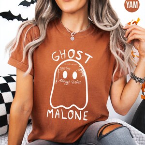 A woman wearing a brown t-shirt with a ghost graphic and the text "GHOST MALONE" is sitting on a couch.