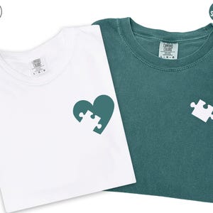 two t-shirts, one white and one green, with a heart-shaped puzzle piece design on the front.