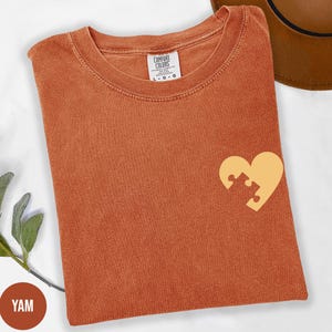 a red t-shirt with a yellow heart-shaped puzzle piece design on the front, placed on a white surface.