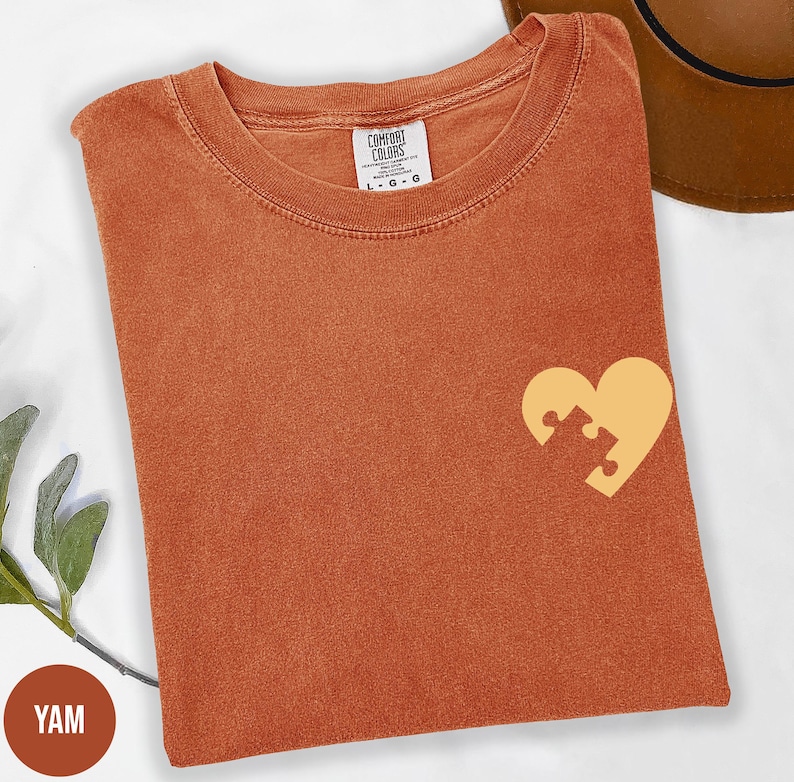 a red t-shirt with a yellow heart-shaped puzzle piece design on the front, placed on a white surface.