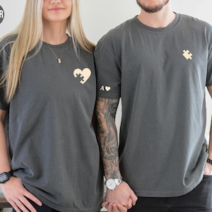 A man and a woman wearing matching gray t-shirts with a heart and puzzle piece design stand together, smiling.