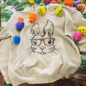 A beige sweatshirt with a black and white illustration of a rabbit wearing glasses, surrounded by colorful Easter eggs on a table.