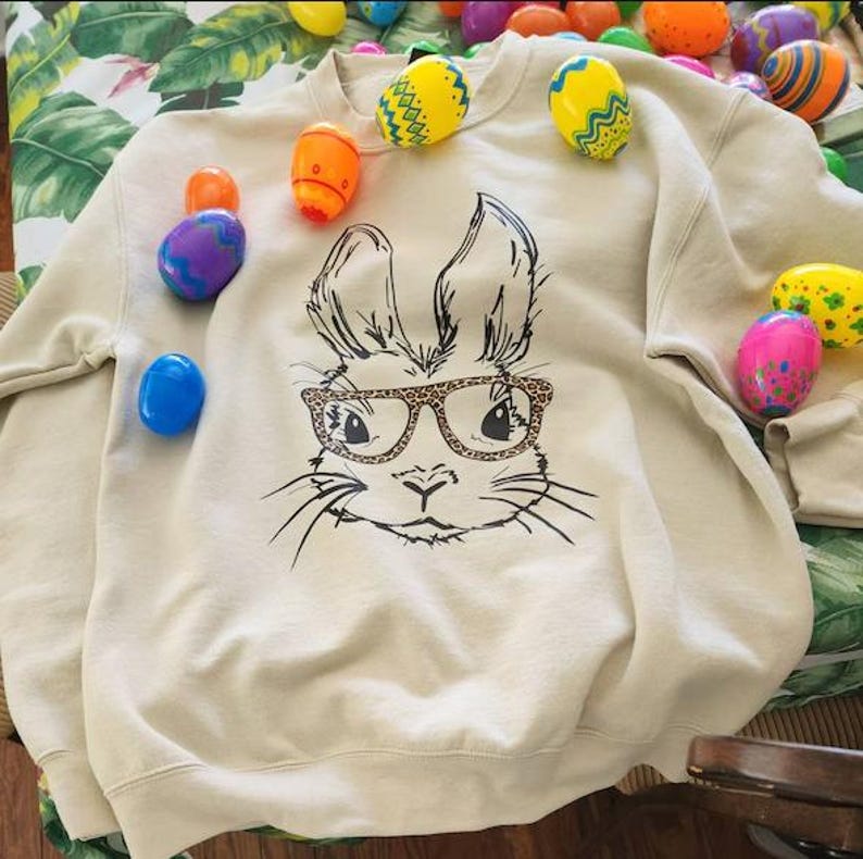 A beige sweatshirt with a black and white illustration of a rabbit wearing glasses, surrounded by colorful Easter eggs on a table.