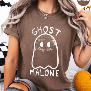 A woman wearing a brown t-shirt with a ghost design and the text "GHOST MALONE" printed on it.