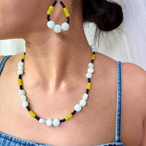 May include: A beaded jewelry set featuring a necklace and matching earrings. The necklace and earrings have a combination of white, yellow, and black beads. The earrings are teardrop-shaped. The jewelry is worn on a person wearing a denim top.