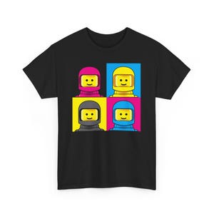 Könnte beinhalten: Schwarzes T-Shirt mit einem Pop-Art-Design von vier Astronautenfiguren. Die Figuren tragen rosa, gelbe, graue und blaue Helme, jeweils mit einem lächelnden Gesicht. Jeder Astronaut hat einen andersfarbigen Hintergrund.