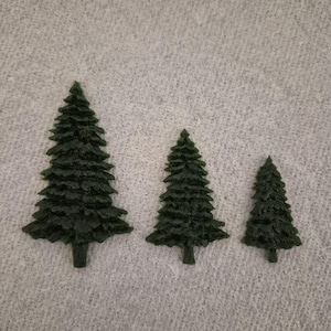 May include: Three dark green miniature Christmas trees of varying sizes. The trees have a detailed, textured appearance, with visible branches and a conical shape. They are arranged in a row against a light gray background.