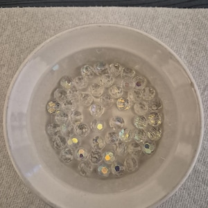 May include: A collection of clear, faceted beads with iridescent highlights, resting in a white ceramic bowl. The beads are round and reflect light, creating a sparkling effect. The bowl is simple and elegant, with a slightly raised rim.